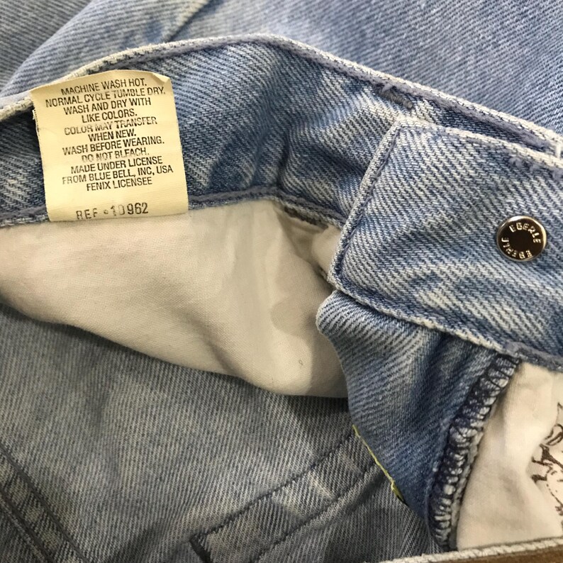 May include: Close-up of light blue denim jeans. The image shows the inside waistband with a tag that reads "MACHINE WASH HOT. NORMAL CYCLE TUMBLE DRY. WASH AND DRY WITH LIKE COLORS..." and a silver button that says "EGERE".