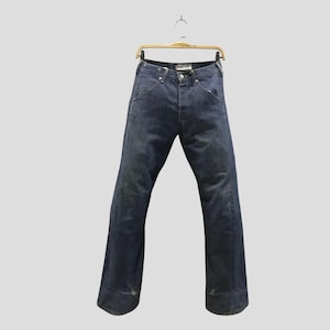 Peut inclure: Un pantalon en jean bleu foncé suspendu à un cintre en métal doré. Le jean présente une taille haute avec une braguette à boutons, deux poches avant et une coupe large qui s'évase légèrement vers l'ourlet inférieur, avec une décoloration subtile.