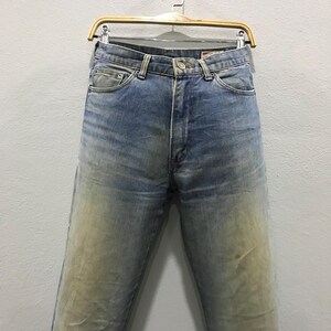 以下が含まれることがあります： 色あせたウォッシュ加工が施された、ライトブルーのデニムジーンズ。クラシックな5ポケットデザイン。木製のハンガーに吊るされています。デニムは、太ももと膝に明るい部分があり、使い古された外観をしています。