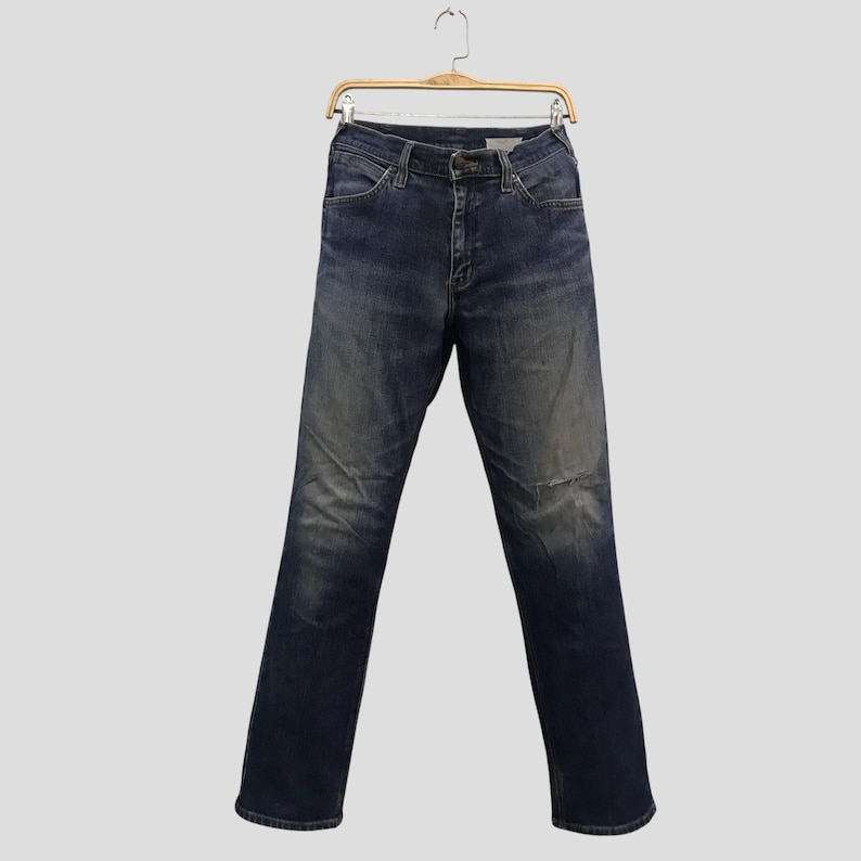 May include: Dark wash denim jeans hanging on a wooden hanger. The jeans have a classic five-pocket design, a button and zip closure, and a faded, worn appearance. The jeans are straight-legged.