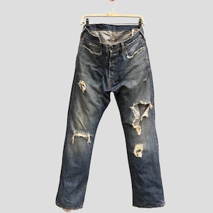 May include: Distressed blue denim jeans with multiple tears and worn areas. The jeans are hanging on a hanger against a light gray background. The jeans have a button fly and classic five-pocket design.