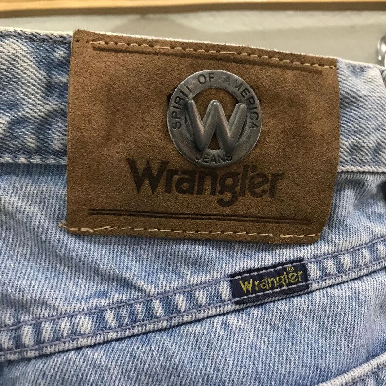 May include: Close-up of light blue denim jeans with a brown leather patch. The patch features a silver emblem with the letter "W" and the words "Spirit of America Jeans" and "Wrangler". A small blue label with "Wrangler" is also visible.