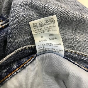 May include: Close-up of denim jeans with a white tag. The tag has Japanese text and symbols indicating care instructions, including a 40-degree wash symbol. The jeans are blue with visible stitching.