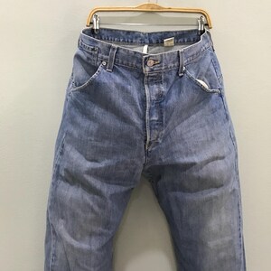 May include: A pair of light blue denim jeans featuring a classic five-pocket design, a button fly closure, and a metal button at the waist. The jeans have a faded wash, visible belt loops, and a visible interior tag at the back waistband.