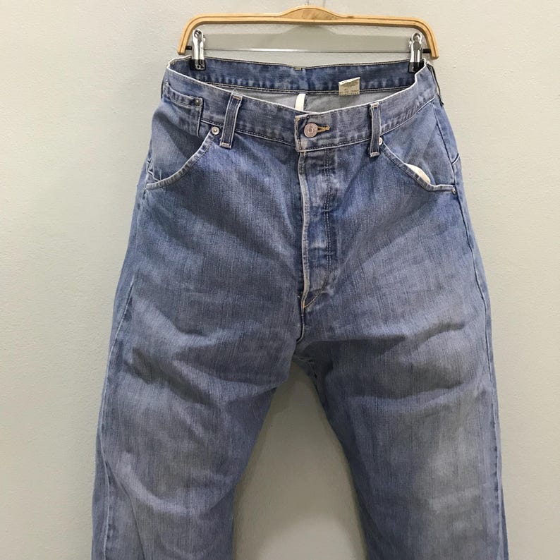 May include: A pair of light blue denim jeans featuring a classic five-pocket design, a button fly closure, and a metal button at the waist. The jeans have a faded wash, visible belt loops, and a visible interior tag at the back waistband.
