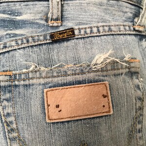 K&ouml;nnte beinhalten: Nahaufnahme der Ges&auml;&szlig;tasche einer hellblauen Jeans. Die Jeans hat einen Used-Look mit ausgefransten Kanten und einem braunen Lederaufn&auml;her. Der Markenname "Wrangler" ist auf einem kleinen schwarzen Etikett zu sehen.