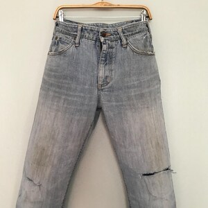 K&ouml;nnte beinhalten: Hellblaue Jeans mit verwaschenem Effekt und Distressed-Details, einschlie&szlig;lich Knie-Rissen und Gebrauchsspuren. Die Jeans h&auml;ngen an einem Holzb&uuml;gel vor einer schlichten wei&szlig;en Wand.