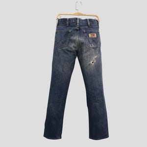Op de afbeelding: Blauwe denim jeans met een versleten look, hangend aan een goudkleurige hanger. De jeans hebben een versleten uitstraling met een scheur op het rechterbeen en een bruine leren patch. Het Wrangler-logo is zichtbaar.