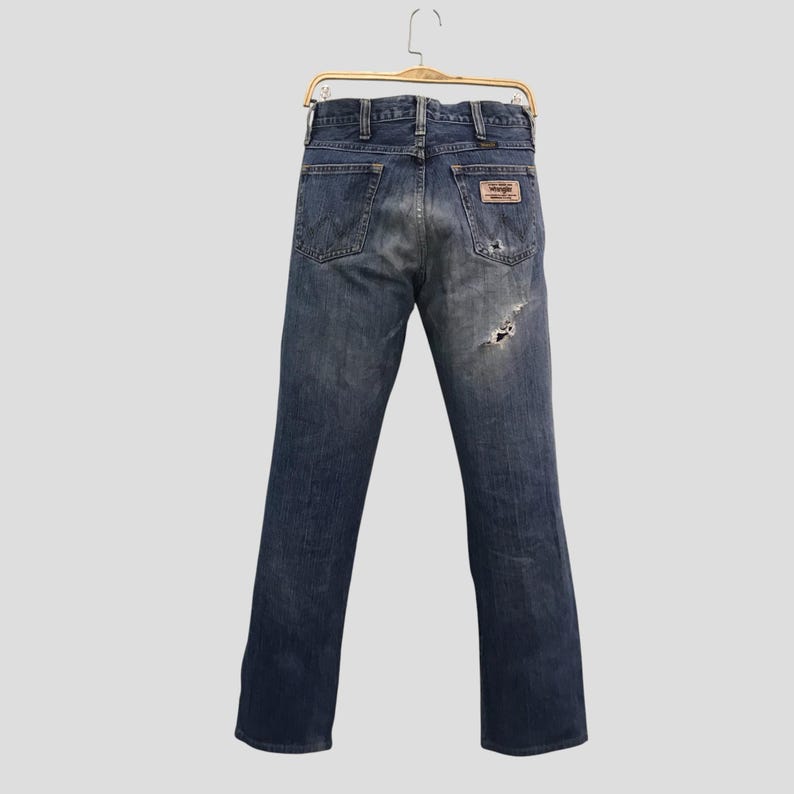 Op de afbeelding: Blauwe denim jeans met een versleten look, hangend aan een goudkleurige hanger. De jeans hebben een versleten uitstraling met een scheur op het rechterbeen en een bruine leren patch. Het Wrangler-logo is zichtbaar.