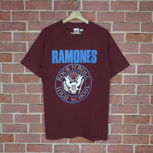 以下が含まれることがあります： 胸にライトブルーのブロック体で「RAMONES」と書かれたマルーン色のTシャツ。その下に、ワシと稲妻のデザインで「ROCK N' ROLL HIGH SCHOOL」と書かれた円形のエンブレムがあります。