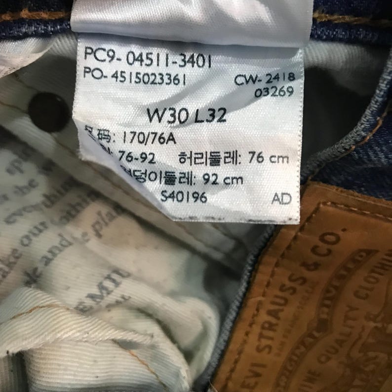 May include: Close-up of a white tag attached to blue denim jeans. The tag displays text including "W30 L32", measurements, and other details. The jeans are from Levi Strauss & Co.