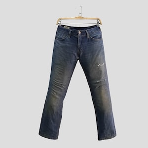 May include: Blue denim jeans with a distressed look, featuring a faded wash and a ripped knee. The jeans are hanging on a wooden hanger, showcasing a classic five-pocket design.