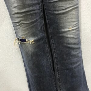 Pu&ograve; includere: Primo piano di jeans blu strappati. I pantaloni in denim hanno un aspetto sbiadito con un ginocchio strappato che rivela una toppa di tessuto blu. I jeans hanno una cucitura verticale al centro.