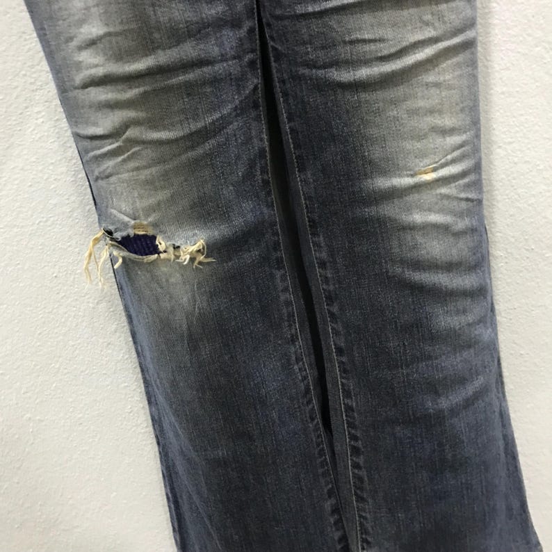 Pu&ograve; includere: Primo piano di jeans blu strappati. I pantaloni in denim hanno un aspetto sbiadito con un ginocchio strappato che rivela una toppa di tessuto blu. I jeans hanno una cucitura verticale al centro.