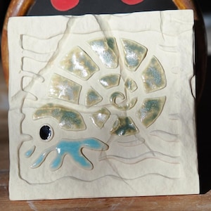 May include: A square off-white ceramic tile featuring a relief carving of an ammonite fossil. The spiral shell has an iridescent finish, while a small blue tentacle-like shape and a black eye are visible at the bottom left of the fossil design.