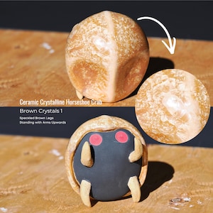 May include: A ceramic crystalline horseshoe crab figurine featuring a glossy brown shell with intricate crystal patterns, a matte black underside, two bright red circular eyes, and four speckled brown legs positioned in an upward-reaching pose.