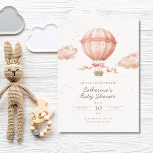May include: A baby shower invitation with a pink hot air balloon illustration, clouds, and the text "Catherine's Baby Shower." A crocheted bunny and wooden toy are on the left. The invitation is on a white wooden surface.
