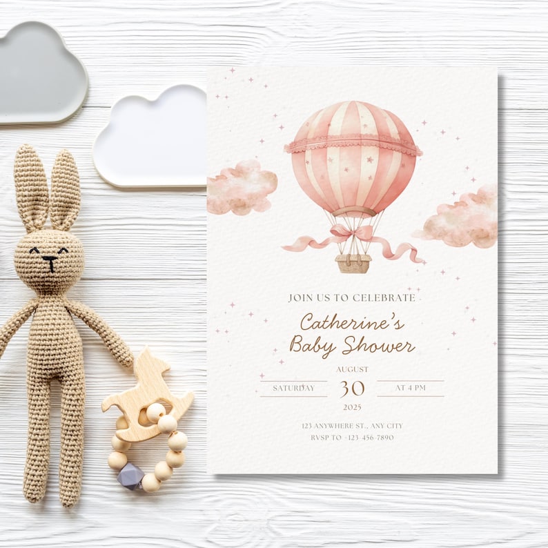 May include: A baby shower invitation with a pink hot air balloon illustration, clouds, and the text "Catherine's Baby Shower." A crocheted bunny and wooden toy are on the left. The invitation is on a white wooden surface.
