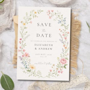 Può includere: Un cartoncino Save the Date rettangolare bianco con una ghirlanda floreale ad acquerello nei toni del rosa, giallo e blu. Il testo recita: SAVE THE DATE TO CELEBRATE THE WEDDING OF ELIZABETH & ANDREW 26th MAY, 2026 City, ST invitation to follow.