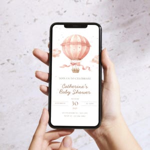 May include: A smartphone displays a baby shower invitation. The invitation features a watercolor illustration of a pink hot air balloon with a basket, ribbons, and clouds. Text reads "Join us to celebrate Catherine's Baby Shower" with date and time.