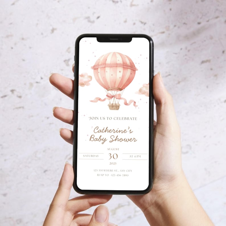 May include: A smartphone displays a baby shower invitation. The invitation features a watercolor illustration of a pink hot air balloon with a basket, ribbons, and clouds. Text reads "Join us to celebrate Catherine's Baby Shower" with date and time.