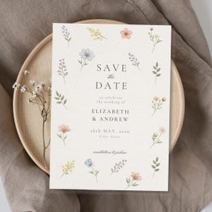 May include: A rectangular Save the Date card featuring a cream-colored textured background with watercolor illustrations of various small wildflowers in shades of blue, yellow, pink, and green, with black text reading Save the Date, Elizabeth & Andrew.