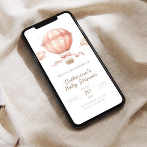 May include: A smartphone displays a baby shower invitation. The invitation features a watercolor illustration of a pink hot air balloon with a basket, ribbons, and clouds. Text reads "Catherine's Baby Shower" and event details.