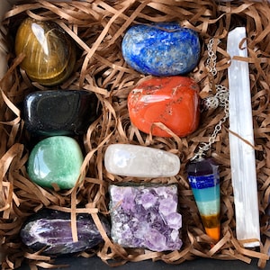May include: A collection of crystals and a pendulum in a brown paper-filled box. The crystals include amethyst, tiger's eye, clear quartz, green aventurine, black obsidian, red jasper, and blue lapis lazuli.