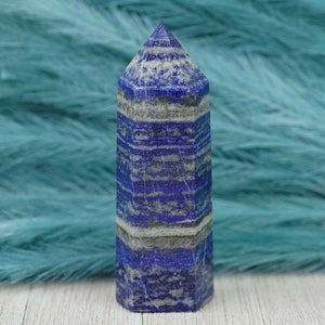 May include: A blue and white striped lapis lazuli crystal point. The crystal is polished and has a pointed tip. It is sitting on a white surface.