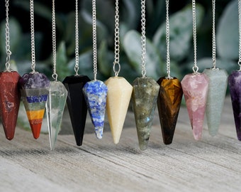 May include: A collection of gemstone pendulums in various colors, including red, purple, black, and blue, each suspended by a silver chain. The pendulums are displayed against a blurred green and white background.