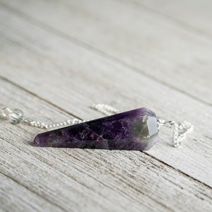May include: Amethyst crystal pendulum with a silver chain. The pendulum is a dark purple color with a pointed tip.