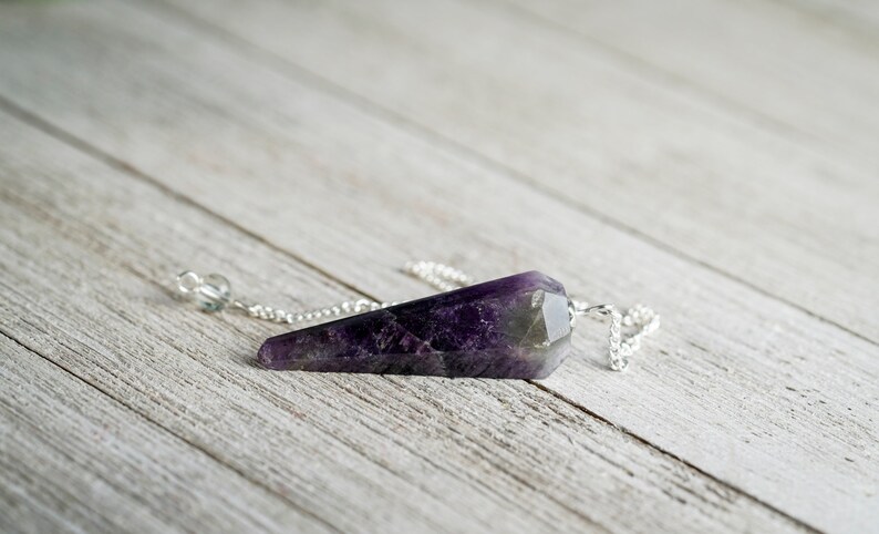 May include: Amethyst crystal pendulum with a silver chain. The pendulum is a dark purple color with a pointed tip.