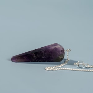 May include: A purple amethyst crystal pendulum with a silver chain. The pendulum is pointed at one end and has a faceted shape.