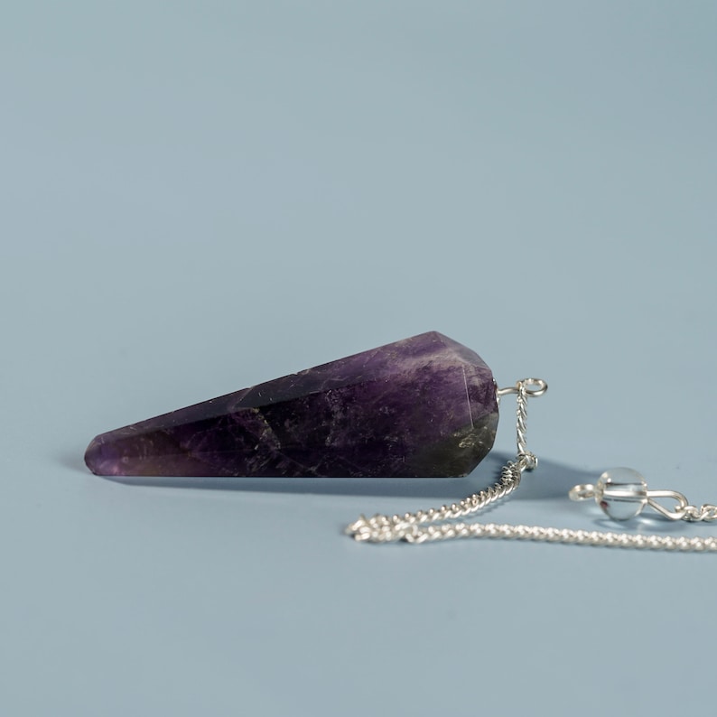 May include: A purple amethyst crystal pendulum with a silver chain. The pendulum is pointed at one end and has a faceted shape.