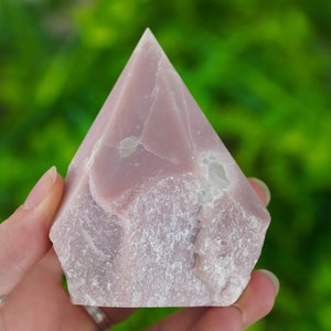 May include: A pink crystal obelisk with a rough, textured surface. The crystal has a pyramid shape with a pointed top and is held against a blurred green background.