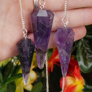 May include: Three amethyst crystal pendulums with silver chains. The pendulums are different sizes and have a pointed tip.