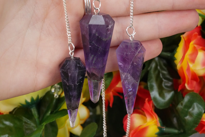 May include: Three amethyst crystal pendulums with silver chains. The pendulums are different sizes and have a pointed tip.