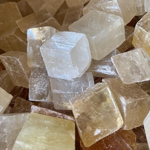 Op de afbeelding: Een close-up van een verzameling ruwe calcietkristallen. De kristallen hebben verschillende vormen en maten, met kleuren variërend van helder tot lichtgeel en bruin. Ze hebben een ruwe, natuurlijke textuur, die de natuurlijke schoonheid van het mineraal benadrukt.