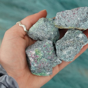 May include: A collection of five rough, gray, green, and red gemstone crystals. The crystals are in the palm of a hand.