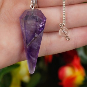 May include: Amethyst crystal pendulum with a silver chain and a clear crystal bead. The pendulum is a deep purple color and has a pointed tip.