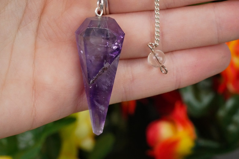 May include: Amethyst crystal pendulum with a silver chain and a clear crystal bead. The pendulum is a deep purple color and has a pointed tip.