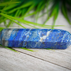 May include: A deep blue lapis lazuli crystal point, also known as a wand, with white veins and a faceted shape. The crystal is lying on a light wood surface with green foliage in the background.