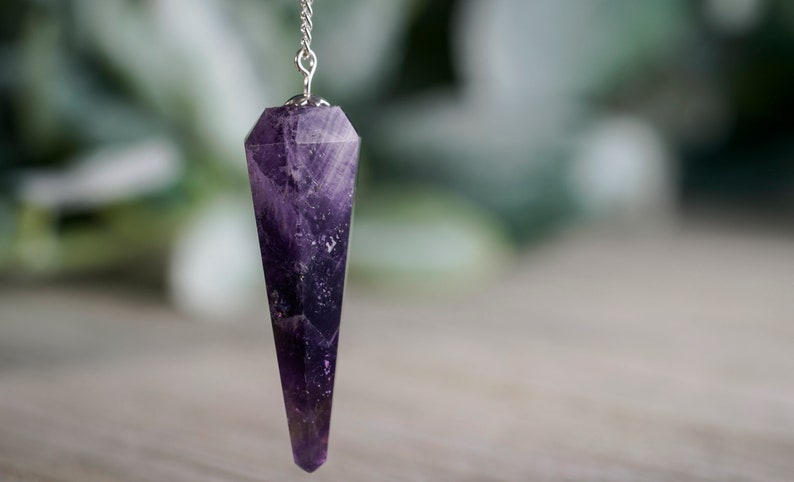 May include: A purple amethyst crystal pendulum on a silver chain. The pendulum is pointed and has a faceted design.