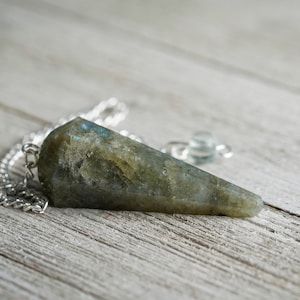 Op de afbeelding: Een gefacetteerde, driehoekige pendel van grijsgroene steen met blauwe en iriserende flitsen, hangend aan een zilveren ketting. De pendel rust op een lichtgrijze houten ondergrond. Een kleine, heldere kraal is aan de ketting bevestigd.