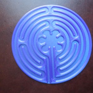 Puede incluir: Un diseño de laberinto circular azul. El laberinto presenta caminos concéntricos que conducen a un patrón central en forma de flor. El diseño se encuentra sobre un fondo marrón oscuro.