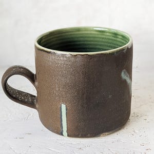 Könnte beinhalten: Handgefertigte Keramik-Tasse mit dunkelbrauner Außenseite und grünem Innenraum. Die Tasse hat eine strukturierte Oberfläche und einen Henkel. Ein vertikaler hellgrüner Streifen ist sichtbar. Geeignet für Kaffee, Tee oder andere Getränke.