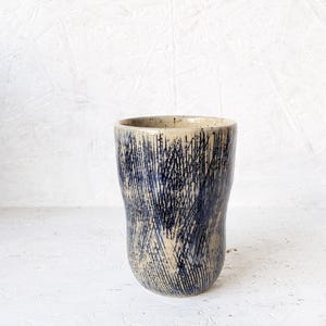 Puede incluir: Un vaso de cerámica hecho a mano con un diseño único. El vaso de color beige presenta un patrón de líneas oscuras, azules y verticales, similares a rasguños. La taza tiene una forma ligeramente irregular y un acabado liso y brillante. Fondo blanco texturizado.