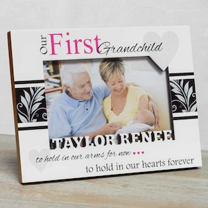 May include: White picture frame with a black and white floral design. The frame has a heart cutout at the top right corner and the text "Our First Grandchild" in pink and black lettering. The text "TAYLOR RENEE" is below the photo opening. The text "to hold in our arms for now... to hold in our hearts forever" is at the bottom of the frame.