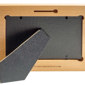 May include: Back view of a wooden picture frame with a black backing and easel. The frame is made of light-colored wood and has a rectangular shape. The easel is made of a dark gray material and is angled to support the frame.