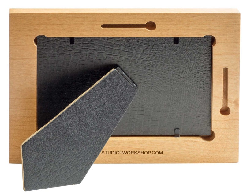 May include: Back view of a wooden picture frame with a black backing and easel. The frame is made of light-colored wood and has a rectangular shape. The easel is made of a dark gray material and is angled to support the frame.